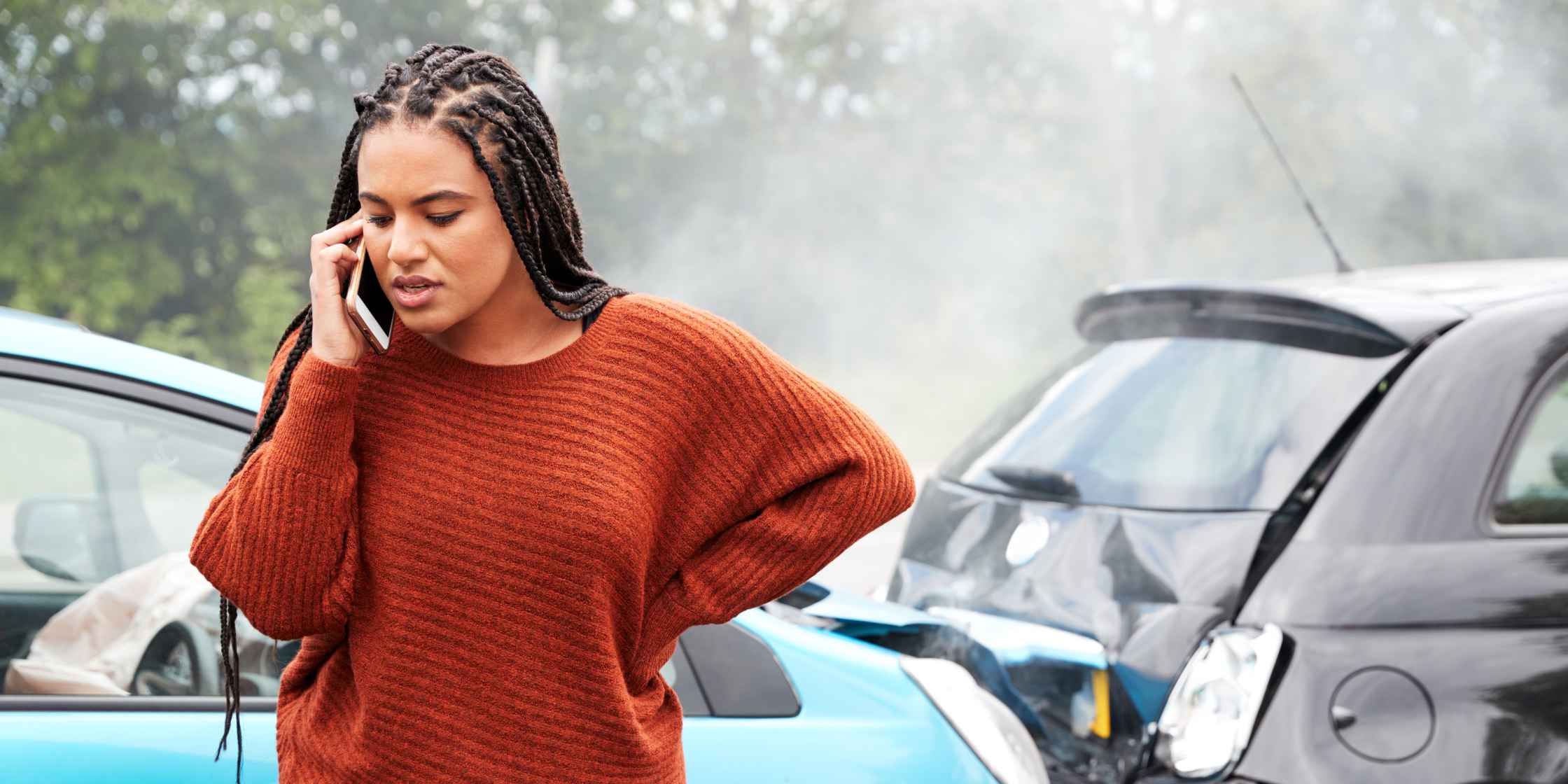 personal-injury-lawyer Distressed woman with braids in a rust sweater urgently calling a personal injury lawyer following a car crash on a foggy day.